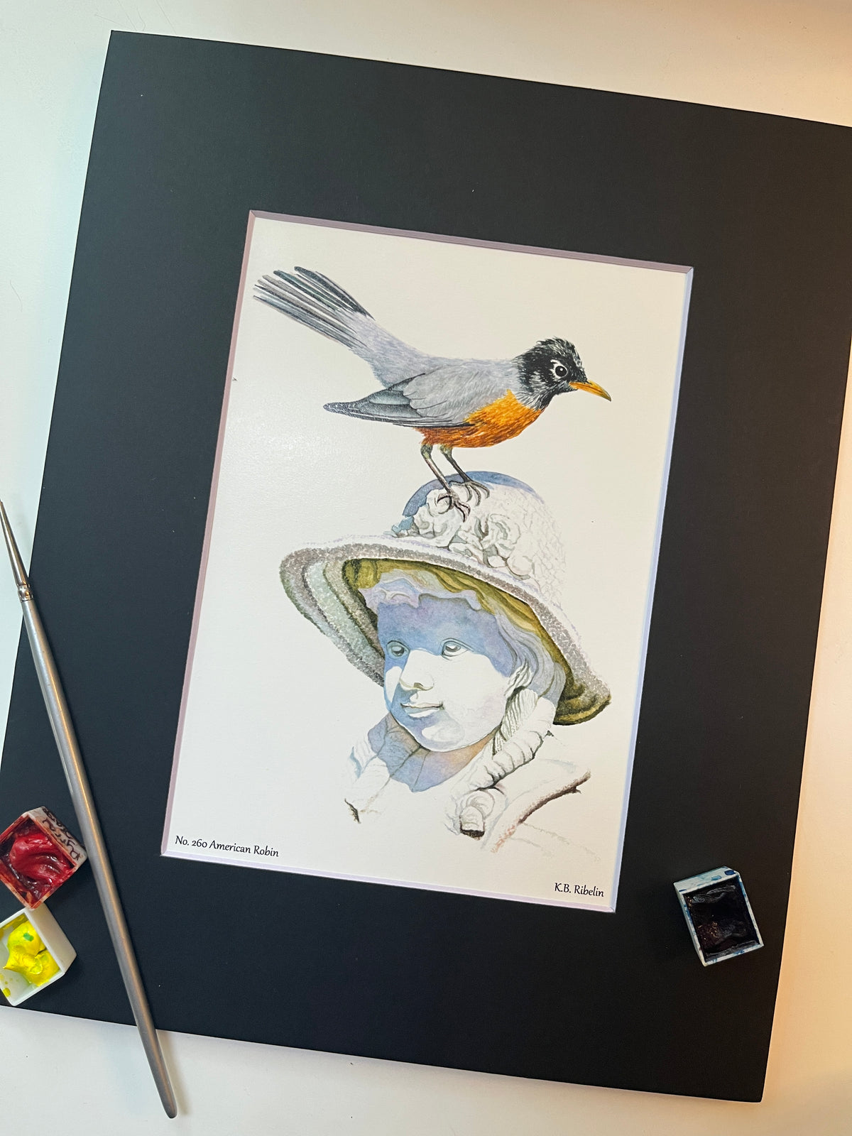 Giclee print of a american robin perched on sculpted girl’s hat displayed in a black mat.