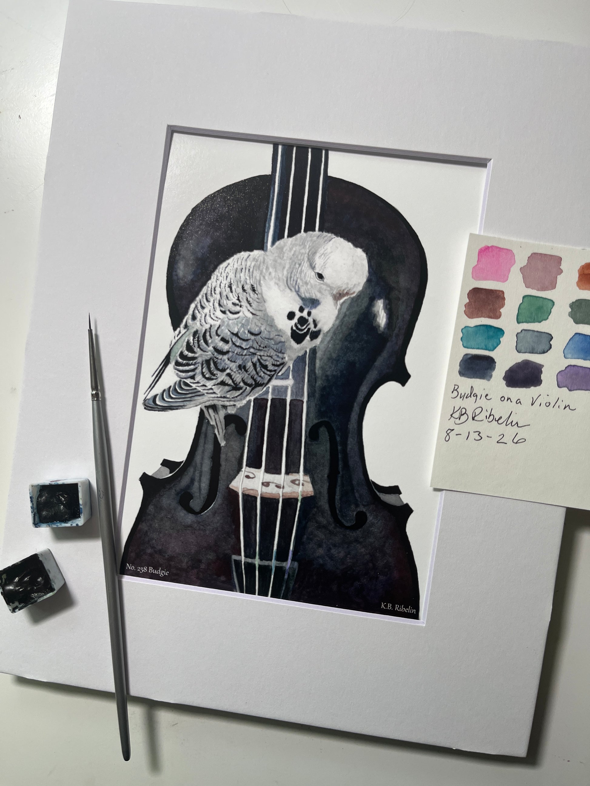 Giclee print of a soft blue‑green budgie perched on a violin watercolor displayed in a white mat.
