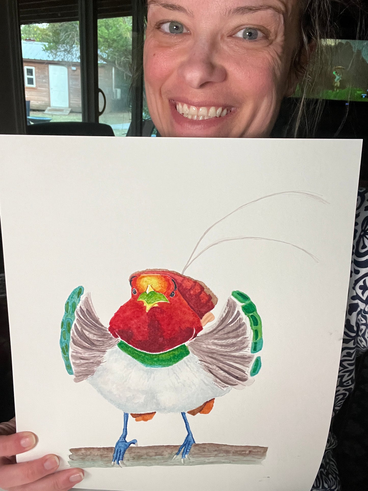 Artist holding the original painting of a king bird-of-paradise perched on branch with red plumage, green collar, and long tail wires, showing the artwork clearly.
