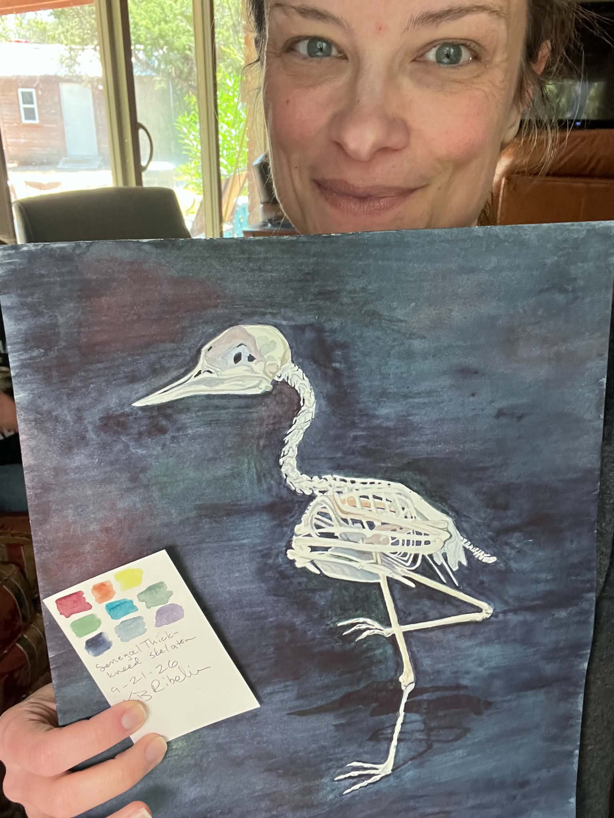 Artist holding the original painting of a senegal thick‑knee bird skeleton study on dark background, showing the artwork clearly.