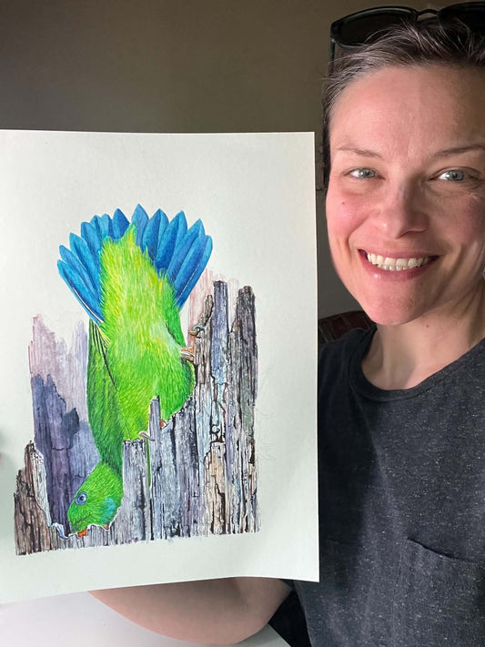 Artist holding the original painting of a vernal hanging parrot perched upside down on weathered wood, showing the artwork clearly.